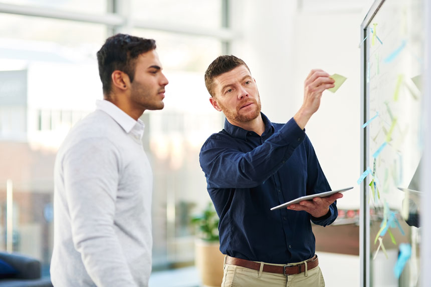 Two men in business casual attire stand in front of a board filled with Post-It notes. The man on the right holds a tablet in one hand and a Post-It note in another.