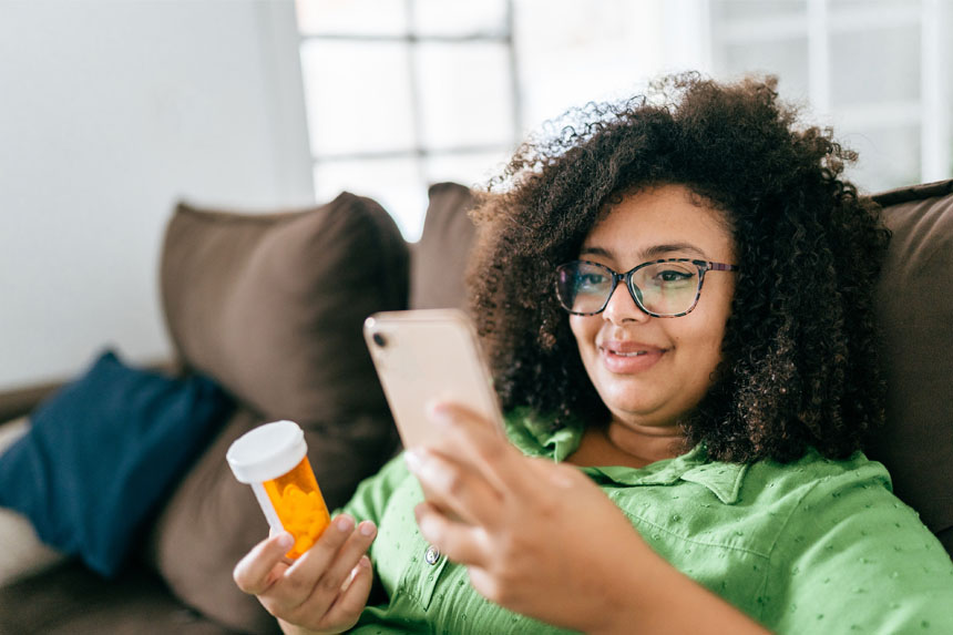 A woman sits on a couch and smiles while looking at her phone in one hand and holding a prescription pill bottle in the other.