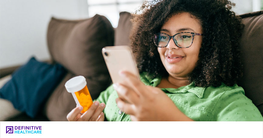 A woman sits on a couch and smiles while looking at her phone in one hand and holding a prescription pill bottle in the other.