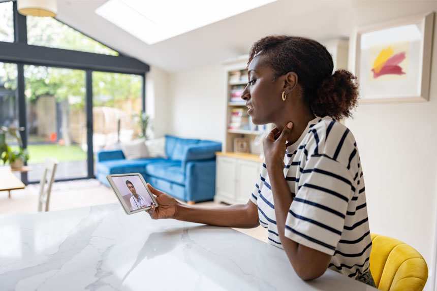 A woman sits at her kitchen counter and holds a tablet computer in one hand and rubs her throat with the other. A doctor, visible on the tablet screen, leads the woman through a telemedicine appointment.