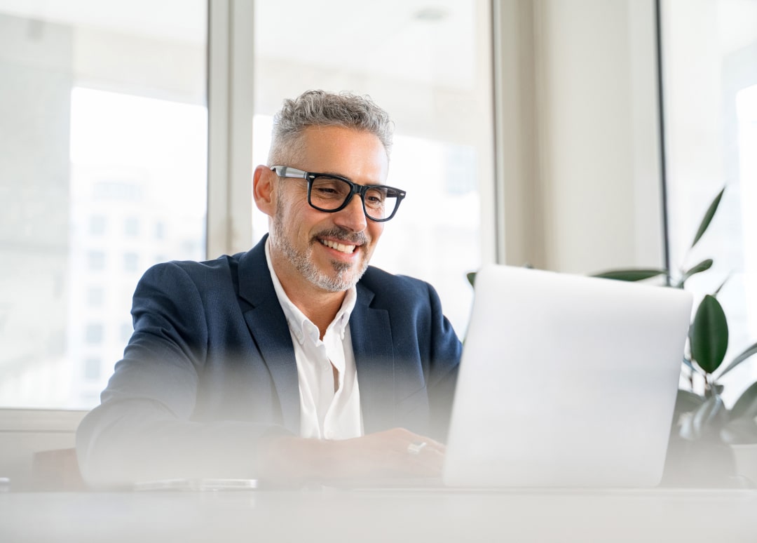 A man in business attire sits at a desk smiling while typing on a laptop.