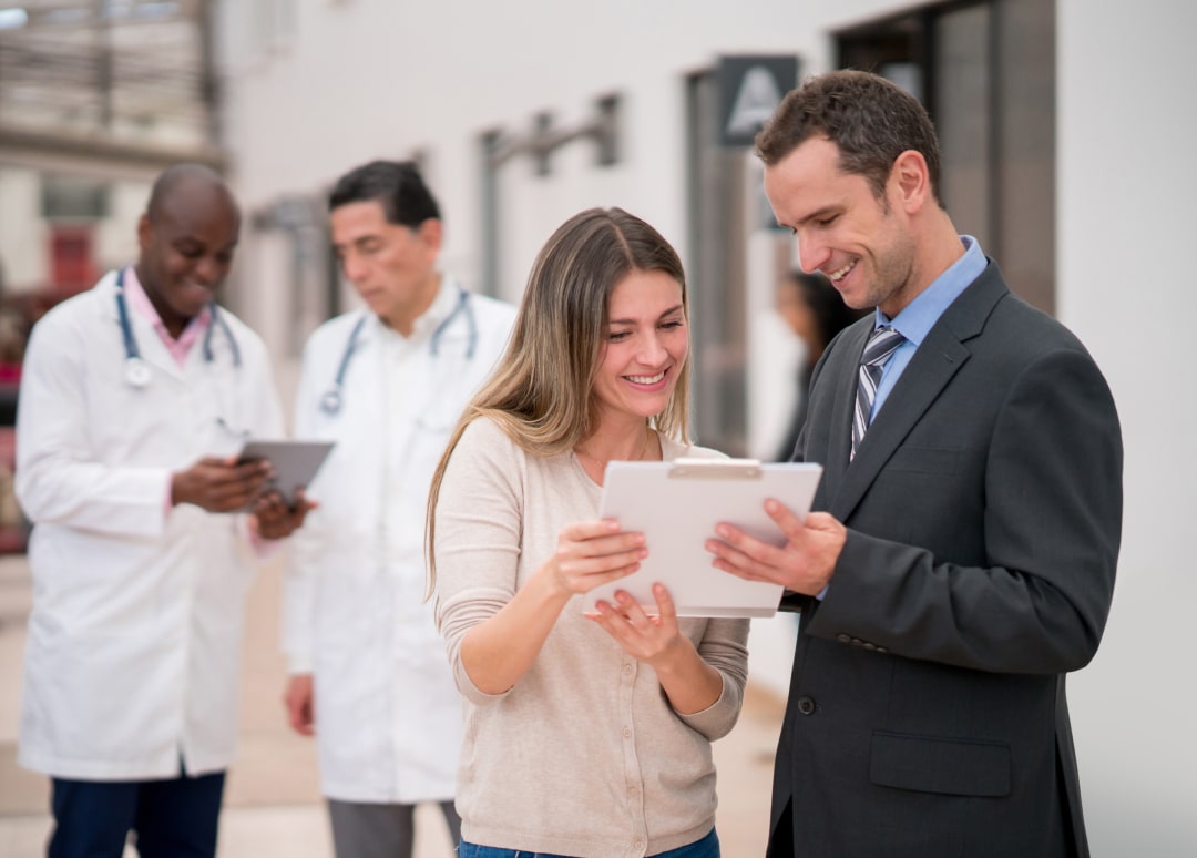 A woman and a man dressed in business attire are smiling while holding and looking down at a clipboard. Two healthcare professionals are standing in the background looking down at a tablet.