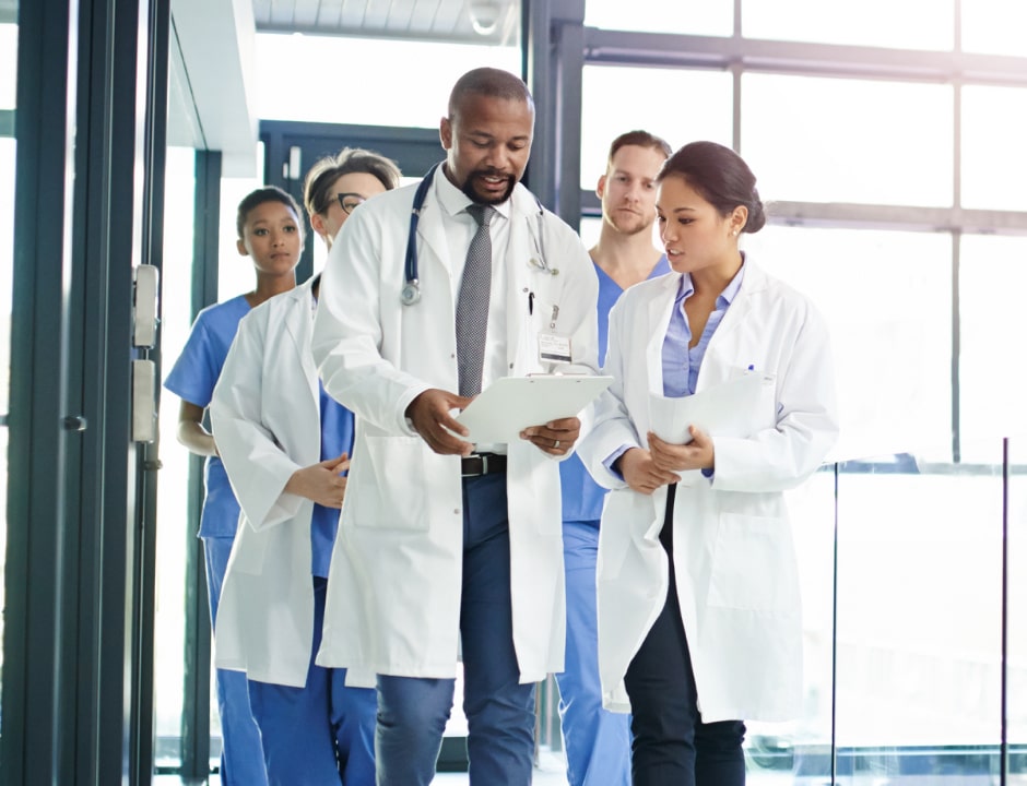 A male doctor is looking down at a clipboard in his hands while walking with a group of healthcare professionals. There is one professional walking next to the doctor holding papers in her hands while also looking down at the clipboard. There are three other healthcare professionals walking behind them.