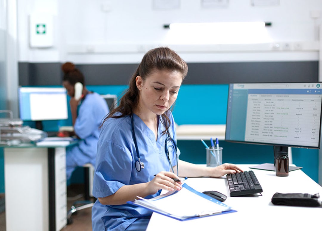 A healthcare professional sits at a desk while working on a computer and looking down at paperwork.