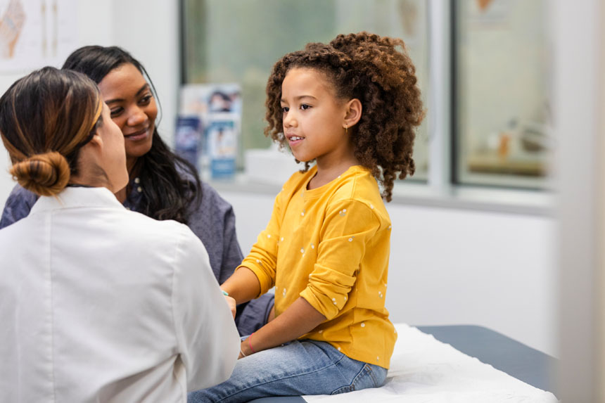 A young girl sits on an examination table in a medical office accompanied by her mother and a female doctor.