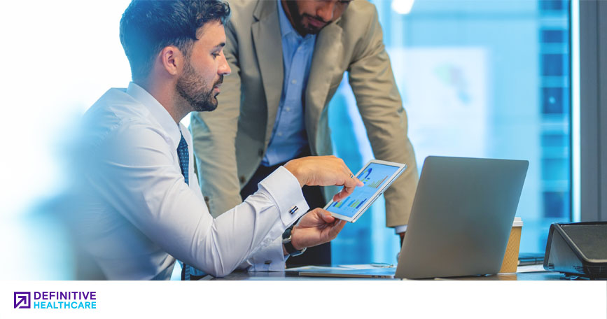 A man in business attire sits in front of a laptop and points to the screen of a tablet computer while another man in business attire leans over to see the screen.