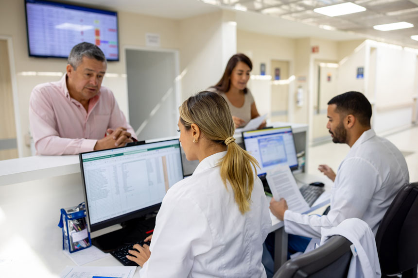 Two medical administrative personnel sit at computer workstations behind a counter in a healthcare facility, helping two patients with billing.