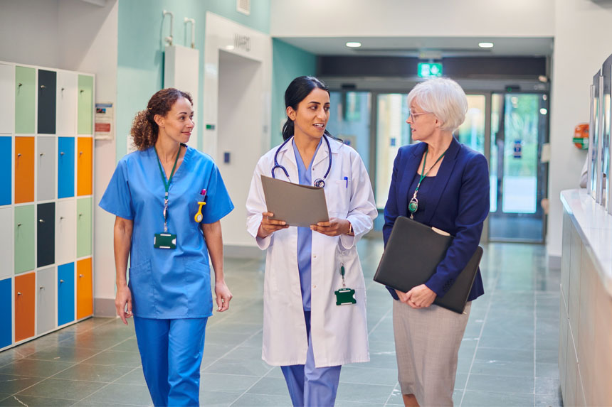 Three female healthcare professionals smile and talk as they walk through a well-lit, modern hospital hallway.