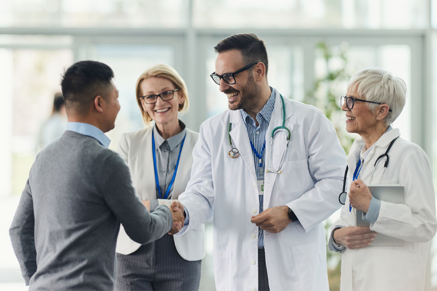 In a well-lit hospital entryway, a person in business attire shakes hands with a smiling physician while two other physicians stand nearby.