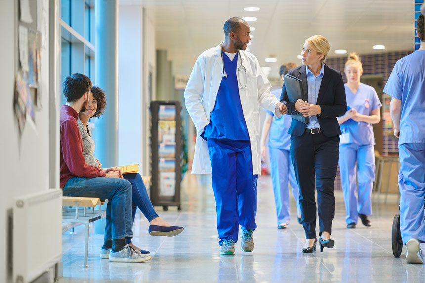 A busy hospital hallway. Two visitors sit and chat on a bench in front of a window, while a doctor talks with a medtech representative. Nurses can be seen walking up and down the hall.