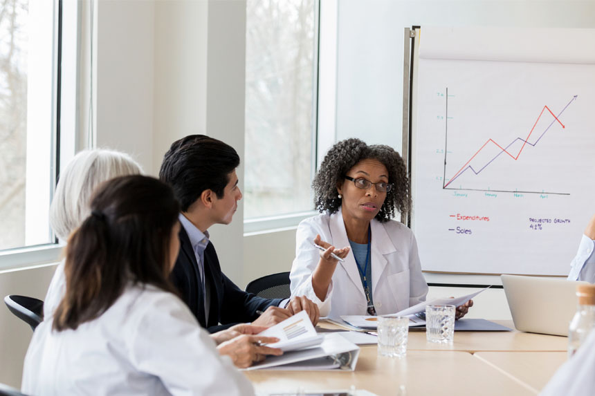 Several healthcare professionals and marketing professionals sit around a conference table and talk while reviewing data. A whiteboard in the background shows a line chart.