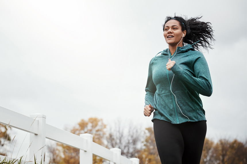 A woman goes on a run next to a white picket fence while wearing a teal hoodie. The sky behind her is overcast.