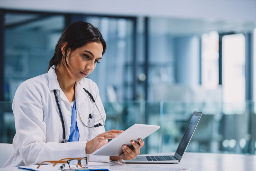 A female healthcare professional examines a tablet while sitting at a desk in a well-lit room.