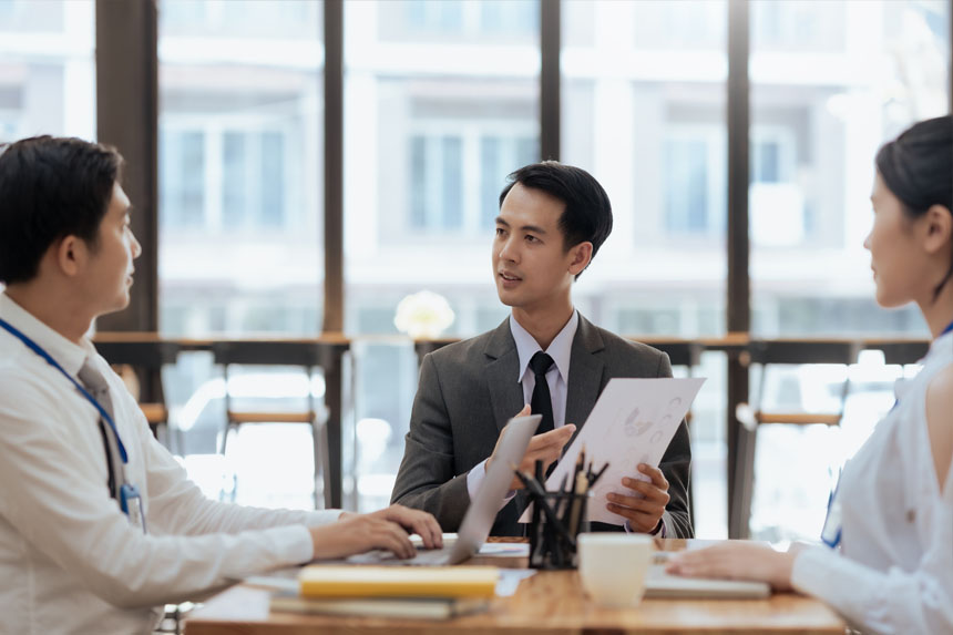 Three people in business attire sit at a conference table in well-lit room, discussing the contents of a report.