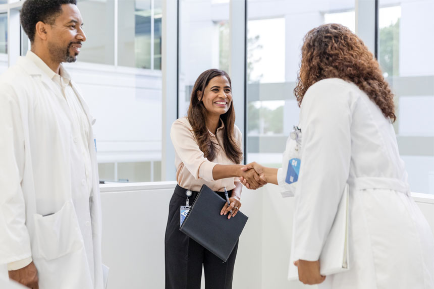 A sales professional shakes hands with a physician while another medical professional looks on.