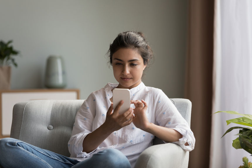 A woman sits on a sofa in her home and operates her smartphone.