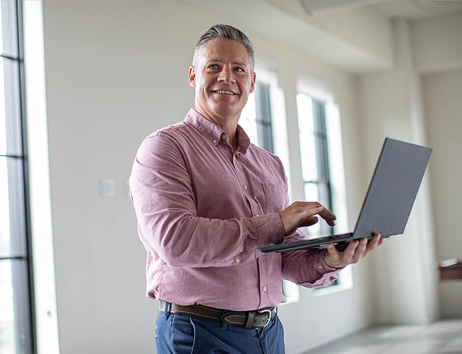 A man in business casual attire is smiling while standing up with a laptop in his hand.