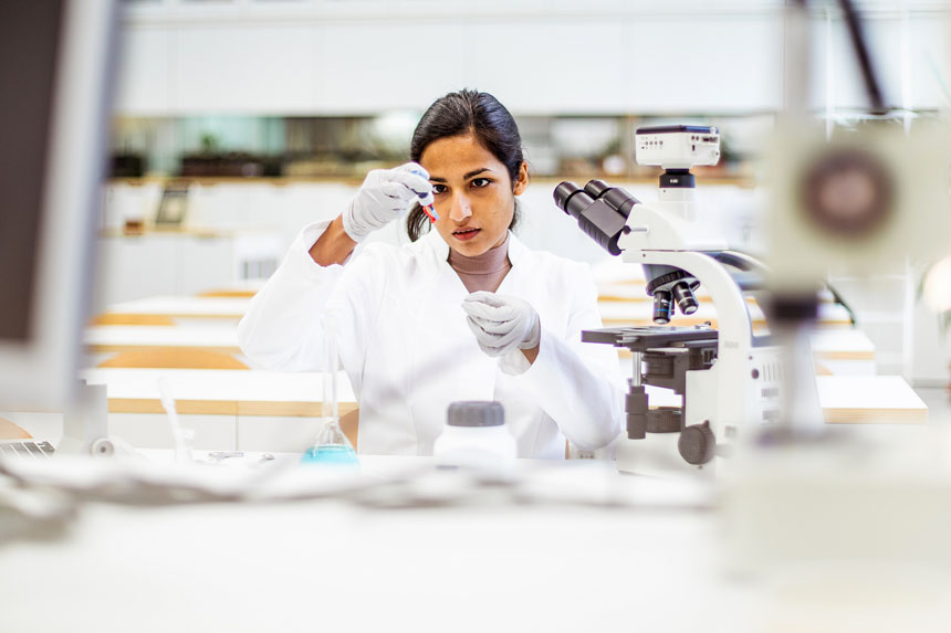 In a well-lit laboratory setting, a woman in a lab coat stands in front of a microscope and examines the contents of a test tube.