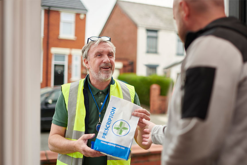 A deliveryman in a hi-viz vest and green polo hands off a prescription bag to a man standing in the open doorway of a suburban home.