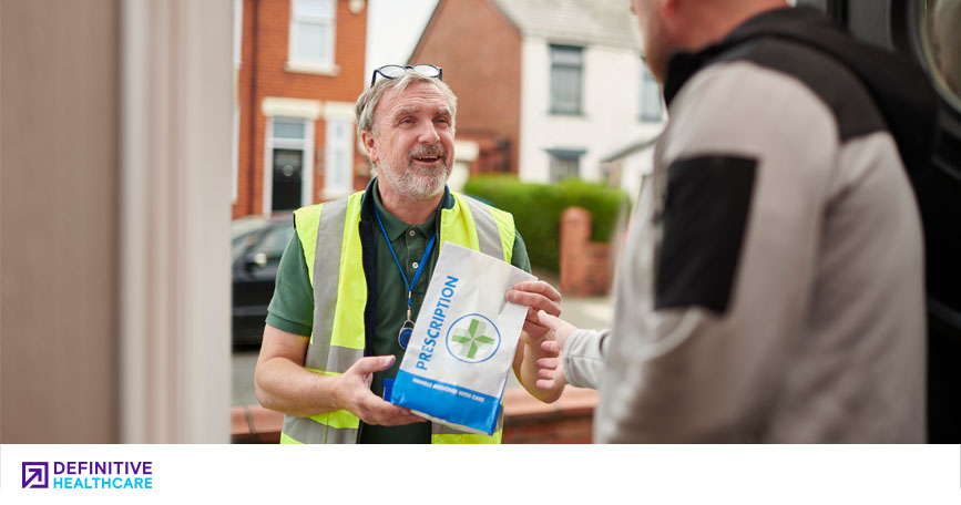 A deliveryman in a hi-viz vest and green polo hands off a prescription bag to a man standing in the open doorway of a suburban home.