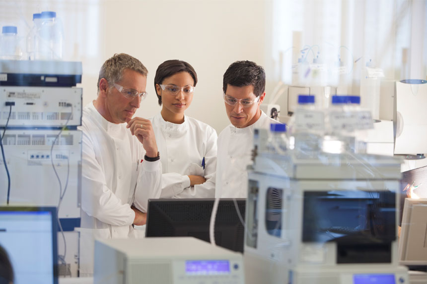 Three researchers wearing white lab attire and safety glasses look at a monitor together.