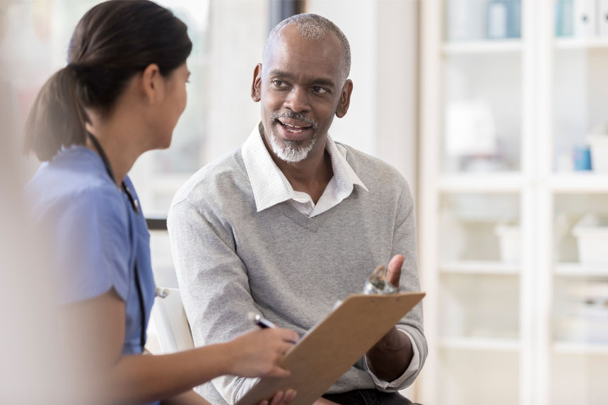 A female healthcare professional talks to a male patient in a well-lit doctor's office.